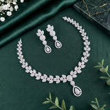 A silver-toned jewelry set featuring a floral leaf-patterned necklace and matching drop earrings, arranged on a dark green satin fabric background with decorative greenery and flowers.