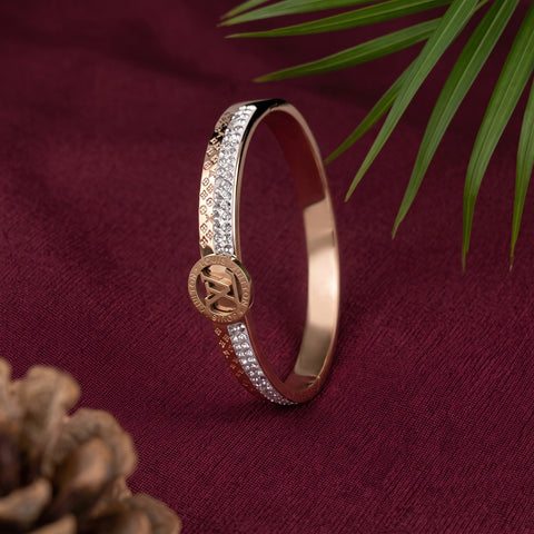 A rose gold hinged bangle bracelet with pavé-set clear stones and a central medallion featuring a monogram, displayed on a deep burgundy fabric background with decorative green leaves and a pinecone.