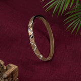 A slim gold-toned bangle bracelet with textured geometric diamond patterns and small clear stone accents, displayed on a deep burgundy fabric background with green palm leaves in the corner.