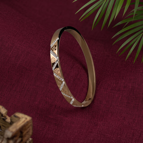 A slim gold-toned bangle bracelet with textured geometric diamond patterns and small clear stone accents, displayed on a deep burgundy fabric background with green palm leaves in the corner.