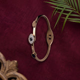 A slim gold-plated bangle bracelet with a central oval cluster of clear stones and a small dark blue bead accent, displayed on a deep burgundy textured fabric background with green palm leaves.