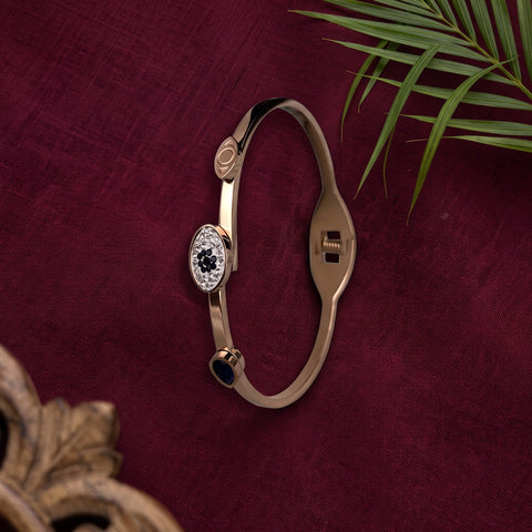 A slim gold-plated bangle bracelet with a central oval cluster of clear stones and a small dark blue bead accent, displayed on a deep burgundy textured fabric background with green palm leaves.