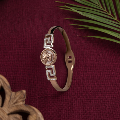 A rose gold bangle bracelet with a central lion emblem and Greek key motifs, displayed on a textured burgundy fabric background with decorative props.
