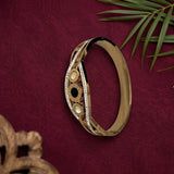 A gold-toned bangle bracelet with intricate circular and oval inlay details, displayed on a textured dark red fabric background with green palm leaves and a decorative wooden object in the corners.
