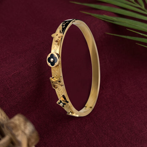A gold-plated bangle bracelet with black enamel floral and geometric details, displayed on a deep burgundy fabric background with green leaves in the corner.