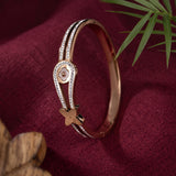 A rose gold bangle bracelet with clear crystal accents and a small flower charm, displayed on a textured dark red fabric background with a green palm leaf.