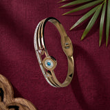 A gold-plated bangle bracelet with a central circular evil eye motif featuring a blue gemstone and mother-of-pearl details, accented with small clear crystals, displayed on a textured burgundy fabric background with a green palm leaf and wooden prop.