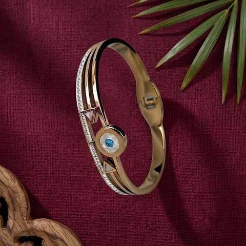 A gold-plated bangle bracelet with a central circular evil eye motif featuring a blue gemstone and mother-of-pearl details, accented with small clear crystals, displayed on a textured burgundy fabric background with a green palm leaf and wooden prop.