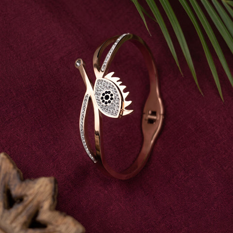 A rose gold hinged cuff bracelet with a stylized eye and leaf design, embellished with clear crystals, displayed on a deep burgundy fabric background with decorative green leaves and wood in the corners.