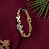 A gold-plated bangle bracelet with three bezel-set clear crystals, displayed on a textured dark red fabric background with decorative green leaves.