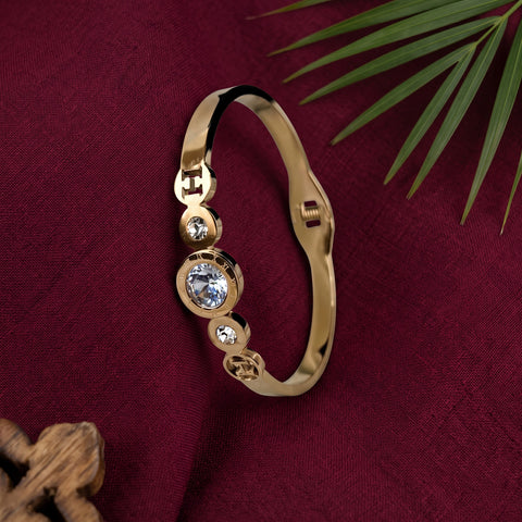 A gold-plated bangle bracelet with three bezel-set clear crystals, displayed on a textured dark red fabric background with decorative green leaves.