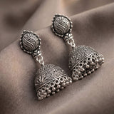 A pair of ornate silver-toned jhumka earrings with intricate filigree detailing and dangling bead chains, displayed on draped taupe fabric.