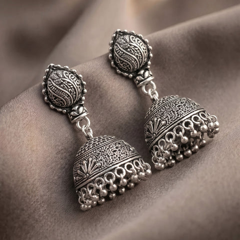 A pair of ornate silver-toned jhumka earrings with intricate filigree detailing and dangling bead chains, displayed on draped taupe fabric.