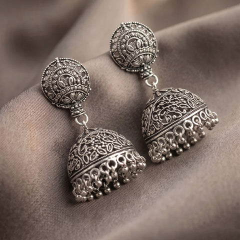 A pair of ornate silver-toned Indian jhumka earrings with intricate filigree detailing and dangling bead tassels, displayed on draped taupe fabric.