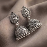 A pair of silver-toned Indian jhumka earrings with intricate floral engravings and dangling bell-shaped drops, displayed on a draped brown fabric background.