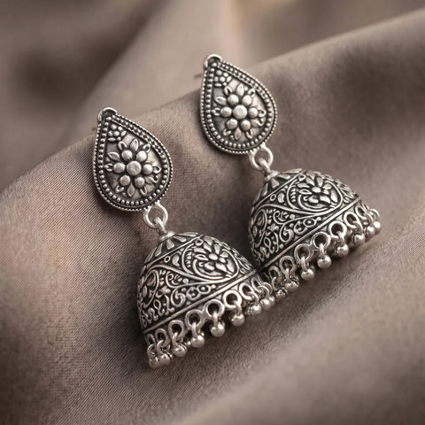 A pair of silver-toned Indian jhumka earrings with intricate floral engravings and dangling bell-shaped drops, displayed on a draped brown fabric background.