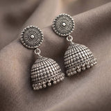 A pair of ornate silver-toned Indian jhumka earrings with intricate circular studs and bell-shaped dangling charms, displayed on draped brown fabric.
