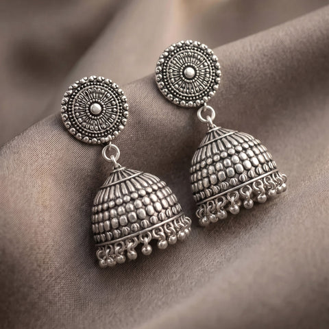 A pair of ornate silver-toned Indian jhumka earrings with intricate circular studs and bell-shaped dangling charms, displayed on draped brown fabric.