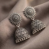A pair of ornate silver-toned Indian jhumka earrings with intricate filigree patterns and dangling bell-shaped drops, displayed on a draped brown fabric background.