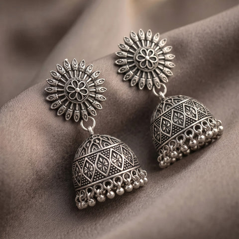 A pair of ornate silver-toned Indian jhumka earrings with intricate filigree patterns and dangling bell-shaped drops, displayed on a draped brown fabric background.