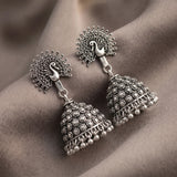 A pair of silver-toned Indian jhumka earrings with peacock head studs and bell-shaped dangling bells, displayed on draped brown fabric.