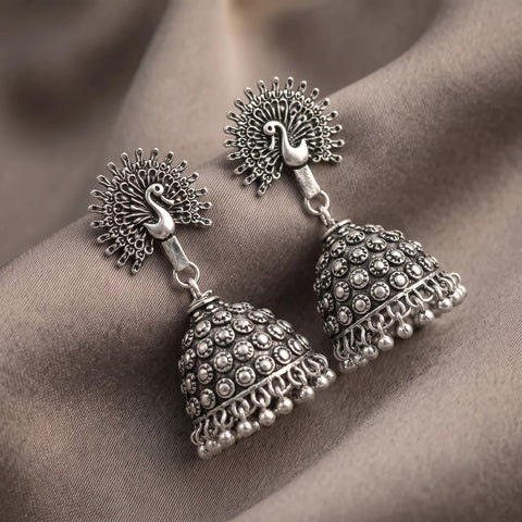 A pair of silver-toned Indian jhumka earrings with peacock head studs and bell-shaped dangling bells, displayed on draped brown fabric.