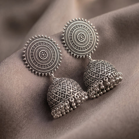 A pair of ornate silver-toned jhumka earrings with intricate circular filigree designs and dangling bell-shaped charms, displayed on draped taupe fabric.