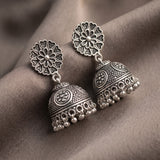 A pair of silver-toned Indian jhumka earrings with intricate filigree floral studs and bell-shaped dangling bells, displayed on a draped brown fabric background.