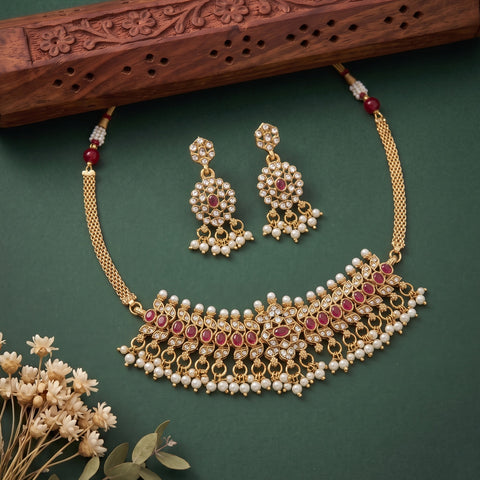 Gold-toned traditional jewelry set featuring a necklace and matching earrings adorned with white pearls and red stones, arranged on a dark green background with decorative dried flowers and a carved wooden prop.
