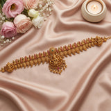 An ornate gold-toned traditional necklace with intricate filigree and small pink gemstone accents, laid flat on draped blush satin fabric with pink roses and a lit candle in the background.