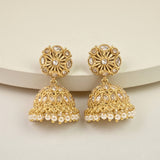 A pair of gold-toned Indian jhumka earrings with clear stones and white faux pearls, displayed on a light beige curved surface.