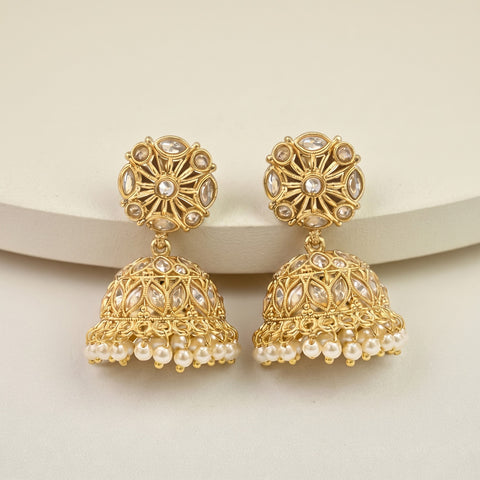 A pair of gold-toned Indian jhumka earrings with clear stones and white faux pearls, displayed on a light beige curved surface.