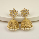 A pair of gold-toned Indian jhumka earrings with floral stud tops set with clear stones and dangling bell-shaped charms embellished with small white pearls, displayed on a light beige surface.