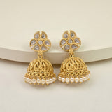 A pair of gold-toned Indian jhumka earrings with white faux-pearl bead borders and teardrop-shaped white stones on the studs, displayed on a light beige surface.