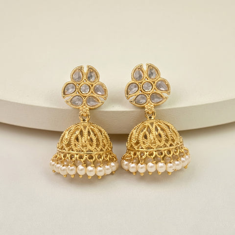 A pair of gold-toned Indian jhumka earrings with white faux-pearl bead borders and teardrop-shaped white stones on the studs, displayed on a light beige surface.