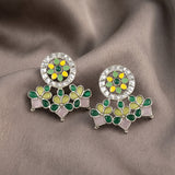 A pair of Indian style Kundan earrings with green and yellow floral motifs, pink stone accents, and a silver crystal-encrusted circular top, displayed on a draped brown fabric background.