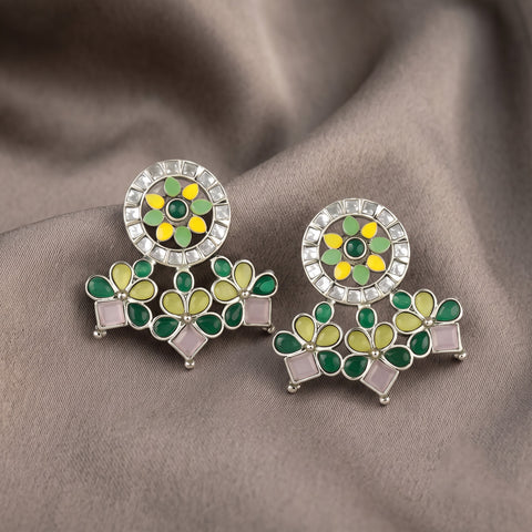 A pair of Indian style Kundan earrings with green and yellow floral motifs, pink stone accents, and a silver crystal-encrusted circular top, displayed on a draped brown fabric background.