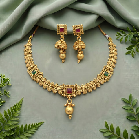 Gold-toned traditional jewelry set with a necklace and matching earrings, featuring red rectangular stones and small green stone accents, arranged on a soft sage-green fabric with decorative green leaves around the edges.