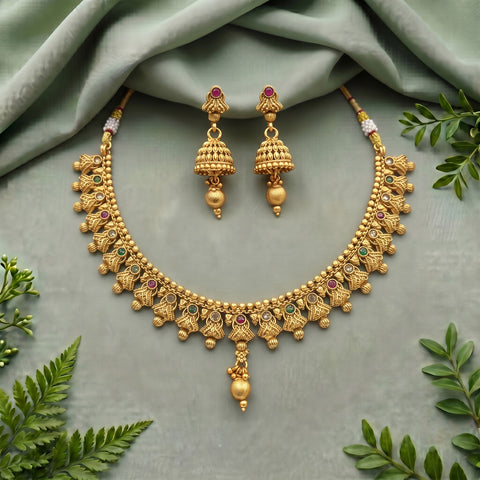 An ornate gold-toned traditional necklace and matching drop earrings laid flat on a soft sage green fabric, with small green and pink gemstone accents and leafy green props around the edges.