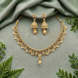 Gold-toned traditional Indian jewelry set featuring a necklace and matching earrings, decorated with gold leaf-shaped discs, small beads, and colorful stone accents, arranged on a draped sage green fabric with green leaves around the edges.