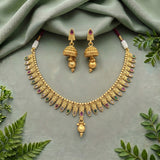 Gold-toned traditional jewelry set with a necklace and matching earrings, decorated with small red and green stones and white pearl-like accents, arranged on a soft sage-green fabric with leafy props around the edges.