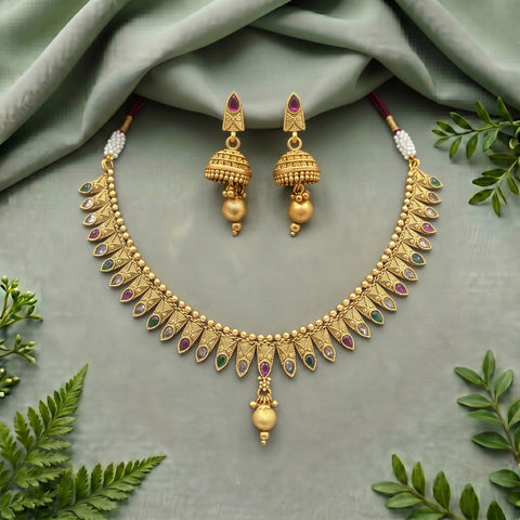 Gold-toned traditional jewelry set with a necklace and matching earrings, decorated with small red and green stones and white pearl-like accents, arranged on a soft sage-green fabric with leafy props around the edges.