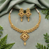 An ornate gold-toned jewelry set featuring a necklace and matching earrings, decorated with red gemstone accents and intricate filigree details, arranged on a draped sage green fabric with decorative green leaves.