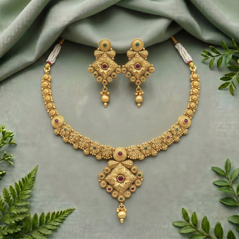 An ornate gold-toned jewelry set featuring a necklace and matching earrings, decorated with red gemstone accents and intricate filigree details, arranged on a draped sage green fabric with decorative green leaves.