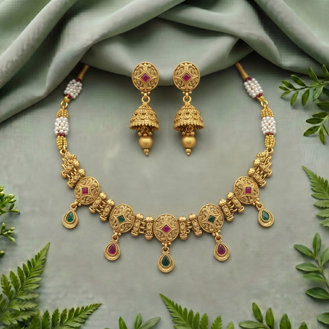 Gold-toned traditional jewelry set with a necklace and matching earrings, decorated with small red and green stones and white pearl-like beads, arranged on a soft sage green fabric with green leaves around it.