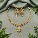 Gold-toned traditional Indian jewelry set featuring a necklace and matching earrings with floral motifs, red and green stone accents, and dangling pearl-like beads, arranged on a draped sage green fabric with decorative green leaves.