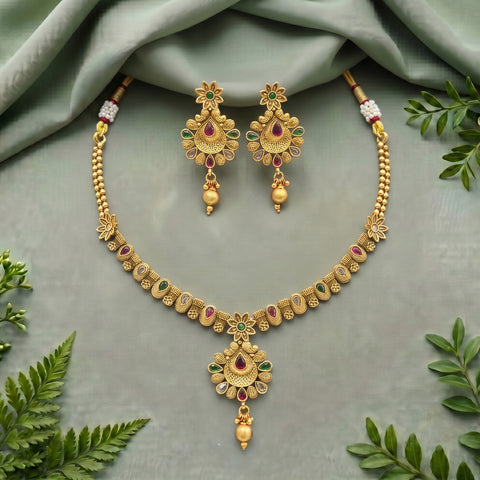 Gold-toned traditional Indian jewelry set featuring a necklace and matching earrings with floral motifs, red and green stone accents, and dangling pearl-like beads, arranged on a draped sage green fabric with decorative green leaves.