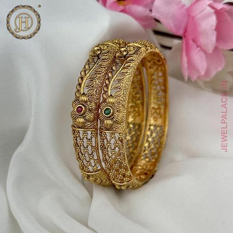 Two gold-plated, floral patterned bangle bracelets with Kemp stone inlays, displayed on a white fabric surface with pink flowers in the background.