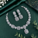 A silver-toned jewelry set featuring a necklace and matching drop earrings with geometric and floral motifs, arranged on a dark green satin fabric background with decorative greenery and a marble slab.