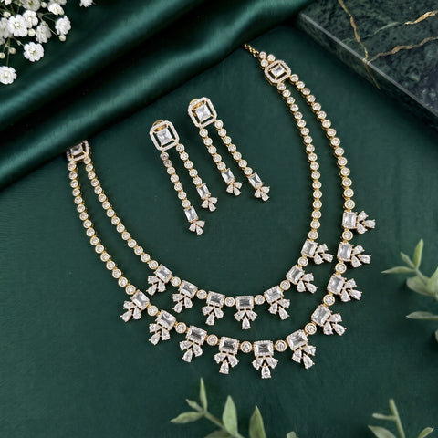 Gold-plated Indian ethnic jewelry set featuring a double-strand necklace, matching drop earrings, and a choker, embellished with clear rectangular and round stones, displayed on a dark green satin fabric background.